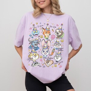 Op de afbeelding: Een lichtpaars T-shirt met cartoonfiguren uit Zootopia, elk met een feesthoedje. Het ontwerp bevat een vos, konijn, luiaard en andere personages, met sterren en bloemmotieven. Het T-shirt heeft een losse pasvorm.