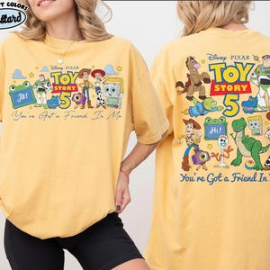 Może przedstawiać: Musztardowa koszulka z grafiką postaci z filmu Disneya Pixara Toy Story. Prz&oacute;d koszulki przedstawia napis "Toy Story 5" i "You've Got a Friend In Me". Tył koszulki r&oacute;wnież ma grafikę Toy Story.