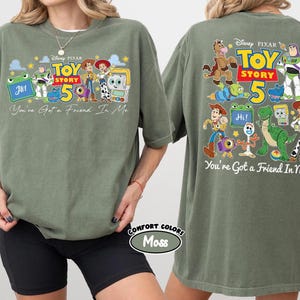 Może przedstawiać: Zielona koszulka w kolorze mchu z grafiką Toy Story i napisem "Toy Story 50". Z przodu koszulki znajduje się napis "You've Got a Friend In Me" oraz żaba z napisem "Hi!". Z tyłu koszulki r&oacute;wnież znajduje się napis "You're Got a Friend In Me".