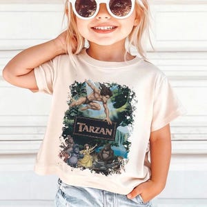 May include: A beige t-shirt featuring a graphic of Tarzan swinging through a jungle scene, with the word "Tarzan" in a dark brown frame. The shirt also depicts various characters from the movie. The shirt is being worn by a person.
