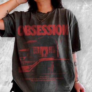 May include: A charcoal grey short-sleeve t-shirt featuring the red text OBSESSION in a bold font. Below the text is a red graphic of a house silhouette with a figure standing in the doorway. Small red dictionary definition text is printed at the bottom.
