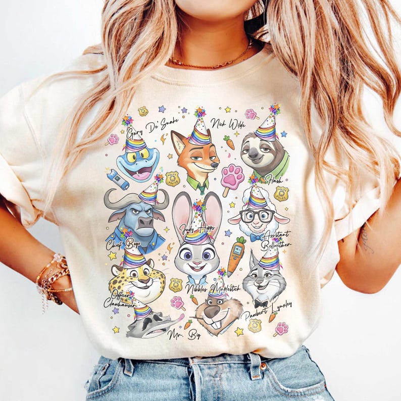 Op de afbeelding: Een cr&egrave;mekleurig T-shirt met stripfiguren, elk met een feesthoedje. De personages zijn onder meer een vos, een luiaard, een konijn en anderen, met namen als Nick Wilde en Judy Hopps. Kleurrijke sterren en feestversieringen omringen de personages.