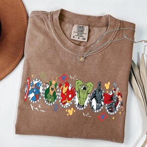 Puede incluir: Una camiseta marr&oacute;n Comfort Colors con un gr&aacute;fico de superh&eacute;roes de Marvel en marcos ovalados. La camiseta tiene cuello redondo y mangas cortas. Un collar plateado con un colgante est&aacute; sobre la camiseta.