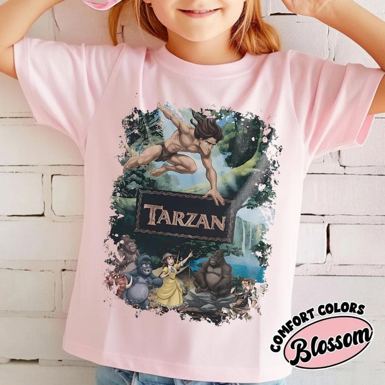May include: A light pink t-shirt featuring a graphic of Tarzan in a jungle setting. The design includes the word "Tarzan" and other characters from the movie. The shirt has short sleeves and a round neckline.