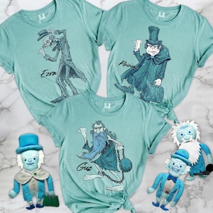 May include: Three light teal t-shirts featuring illustrations of characters labeled Ezra, Phineas, and Gus, accompanied by two plush dolls wearing blue top hats and coats, each with white hair and beards, matching the characters printed on the shirts.
