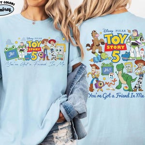 Może przedstawiać: Jasnoniebieska koszulka z nadrukiem z Toy Story, przedstawiającym postacie takie jak Woody i Buzz Lightyear. Widoczne są napisy "TOY STORY 5" i "You've Got a Friend In Me". Koszulka wykonana jest z miękkiego materiału.