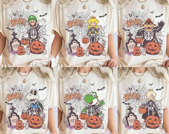 Könnte beinhalten: Ein cremefarbenes T-Shirt mit einem Halloween-Motiv, das ein Spinnennetz, einen Grabstein mit der Aufschrift RIP, Kürbisse, Fledermäuse und eine zentrale Figur in einem Skelettkostüm sowie das Wort BOO in orangefarbenen Buchstaben zeigt.