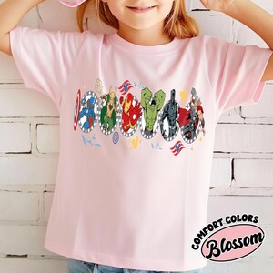 Puede incluir: Camiseta rosa claro con un gr&aacute;fico colorido de superh&eacute;roes en marcos ovalados. La camiseta tiene las palabras "COMFORT COLORS" y "Blossom" en un dise&ntilde;o circular.