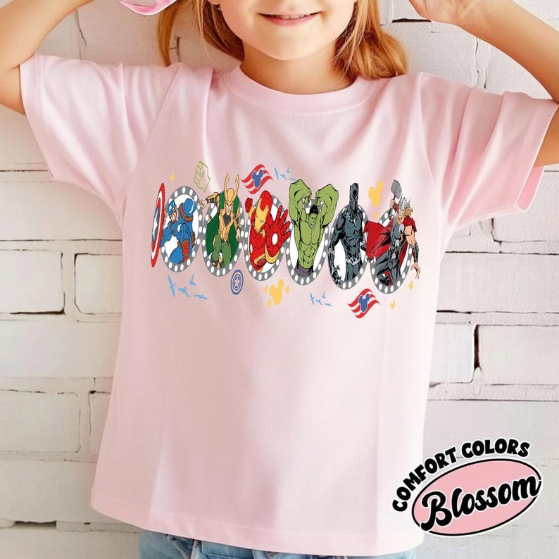 Puede incluir: Camiseta rosa claro con un gr&aacute;fico colorido de superh&eacute;roes en marcos ovalados. La camiseta tiene las palabras "COMFORT COLORS" y "Blossom" en un dise&ntilde;o circular.