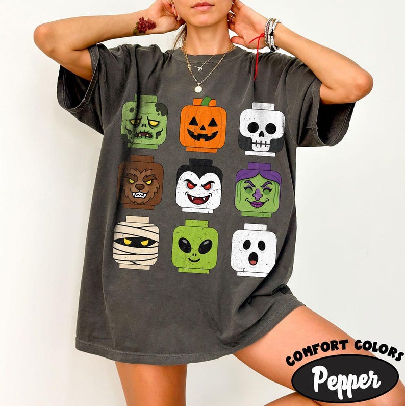 May include: A charcoal gray short-sleeve t-shirt featuring a grid of nine colorful cartoon monster heads resembling toy building block figures, including a zombie, pumpkin, skull, werewolf, vampire, witch, mummy, alien, and ghost character.