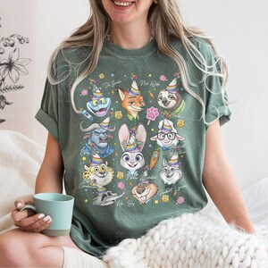 Op de afbeelding: Groen t-shirt met een kleurrijke afbeelding van cartoon dierenfiguren met feesthoeden. Het ontwerp bevat een kikker, vos, luiaard, konijn, schaap, cheetah en andere karakters. De shirt heeft opgerolde mouwen.