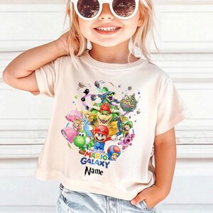 Puede incluir: Camiseta beige claro con un gr&aacute;fico colorido de personajes de Super Mario Galaxy, incluyendo a Mario, Yoshi y Bowser. El dise&ntilde;o tambi&eacute;n presenta el texto "THE SUPER MARIO GALAXY" y "Name".