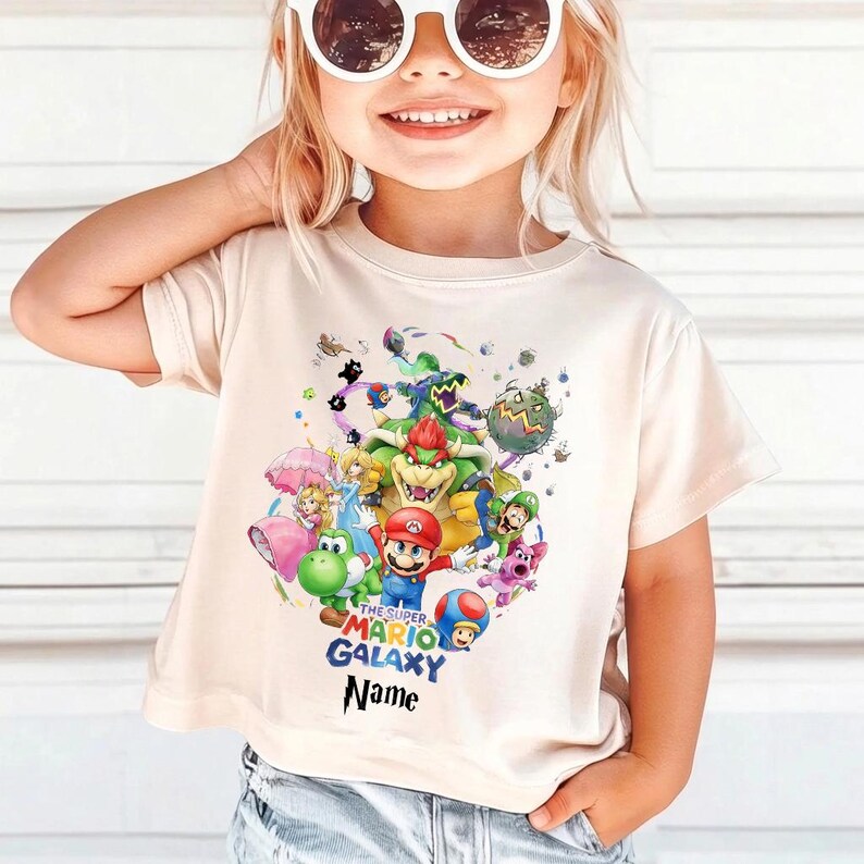 Puede incluir: Camiseta beige claro con un gr&aacute;fico colorido de personajes de Super Mario Galaxy, incluyendo a Mario, Yoshi y Bowser. El dise&ntilde;o tambi&eacute;n presenta el texto "THE SUPER MARIO GALAXY" y "Name".