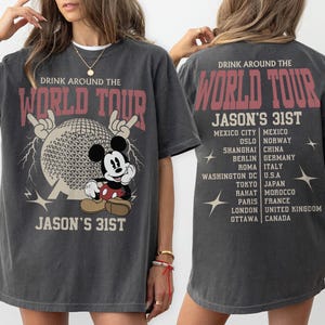 Op de afbeelding: Een antracietgrijs t-shirt met een afbeelding van Mickey Mouse, een geodetische bol en bliksemschichten met de tekst "DRINK AROUND THE WORLD TOUR" en "JASON'S 31ST". De achterkant toont een lijst met steden en landen met dezelfde tekst.