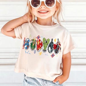 Puede incluir: Una camiseta color crema con un gr&aacute;fico colorido de superh&eacute;roes en marcos ovalados. El dise&ntilde;o incluye al Capit&aacute;n Am&eacute;rica, Thor, Iron Man y Hulk. La camiseta tiene mangas cortas y cuello redondo.