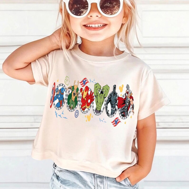 Puede incluir: Una camiseta color crema con un gr&aacute;fico colorido de superh&eacute;roes en marcos ovalados. El dise&ntilde;o incluye al Capit&aacute;n Am&eacute;rica, Thor, Iron Man y Hulk. La camiseta tiene mangas cortas y cuello redondo.