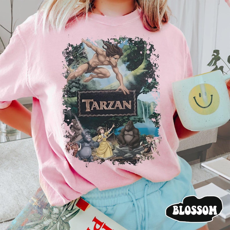 May include: A light pink t-shirt featuring a graphic of Tarzan swinging through a jungle scene, with the word "Tarzan" in a decorative font. The shirt also features other characters from the movie. A light blue mug with a yellow smiley face is held in the image.