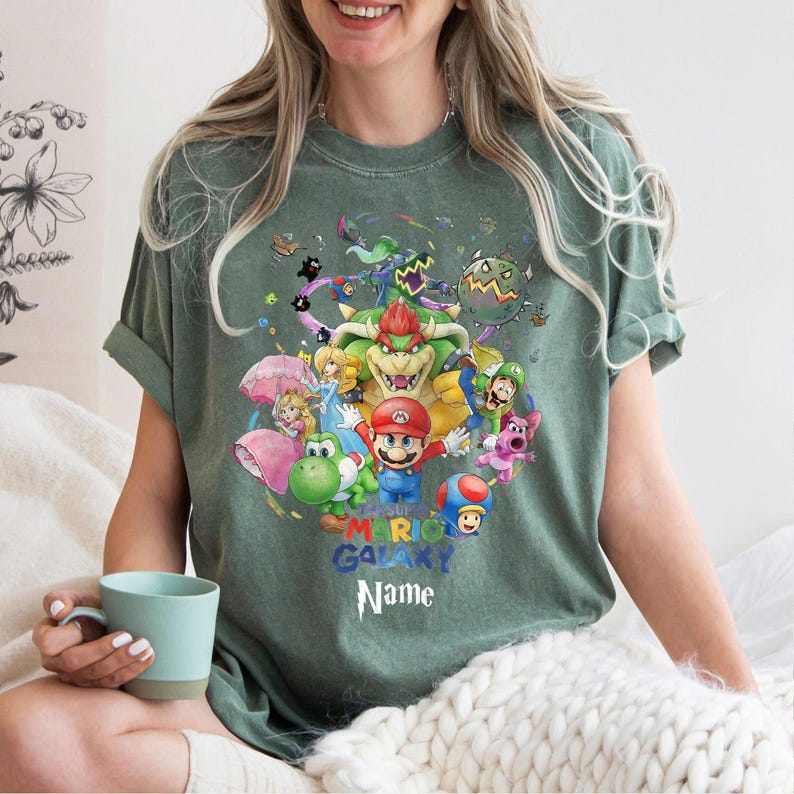 Puede incluir: Camiseta verde con un gr&aacute;fico colorido de Mario y otros personajes del juego Mario Galaxy. El dise&ntilde;o incluye el texto "MARIO GALAXY Name" en azul. La camiseta es de estilo casual y de manga corta.