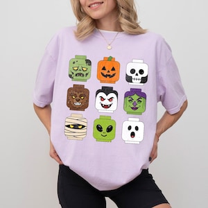May include: A light purple t-shirt featuring a grid of nine stylized block-style character heads. The graphic includes a green zombie, orange pumpkin, white skull, brown werewolf, vampire, green witch, tan mummy, green alien, and a white ghost.