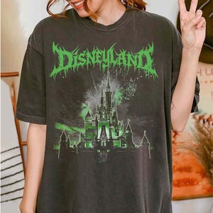 Puede incluir: Camiseta gris oscuro con la palabra "Disneyland" en verde, con texto estilizado sobre un gr&aacute;fico de castillo iluminado en verde con fuegos artificiales. La camiseta es de corte holgado.