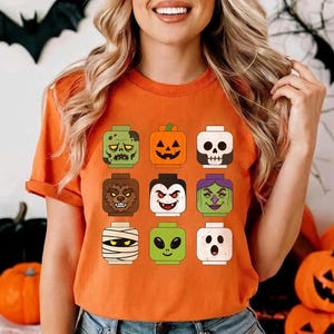 May include: An orange t-shirt featuring a graphic print of nine square-shaped block-style character heads arranged in a grid. The characters include a zombie, a jack-o'-lantern, a skull, a werewolf, a vampire, a witch, a mummy, an alien, and a ghost.