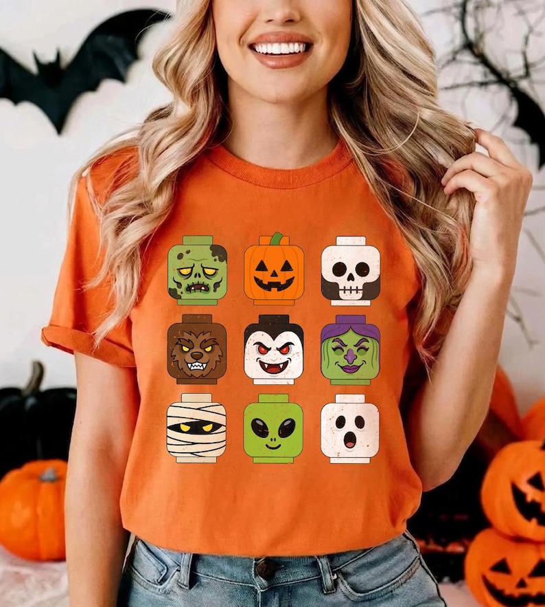 May include: An orange t-shirt featuring a graphic print of nine square-shaped block-style character heads arranged in a grid. The characters include a zombie, a jack-o'-lantern, a skull, a werewolf, a vampire, a witch, a mummy, an alien, and a ghost.