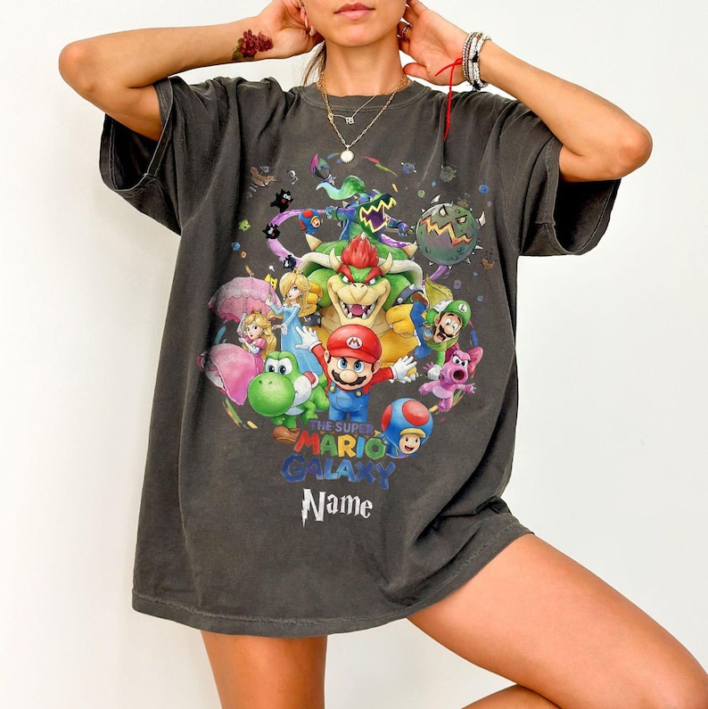 Puede incluir: Camiseta gris oscuro con un gr&aacute;fico colorido de Mario y otros personajes del juego Super Mario Galaxy. El dise&ntilde;o incluye el texto "THE SUPER MARIO GALAXY" y un espacio para un nombre. La camiseta tiene mangas cortas.