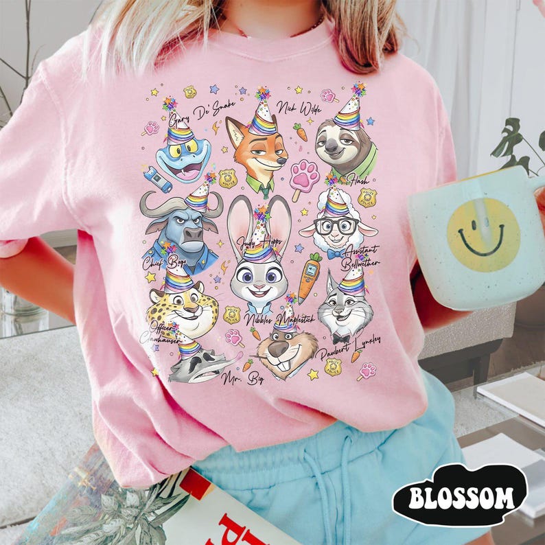 Op de afbeelding: Een lichtroze t-shirt met cartoonfiguren uit Zootopia, elk met een feesthoedje. De personages zijn onder meer Judy Hopps, Nick Wilde en Flash. Op het shirt staat ook het woord "BLOSSOM".