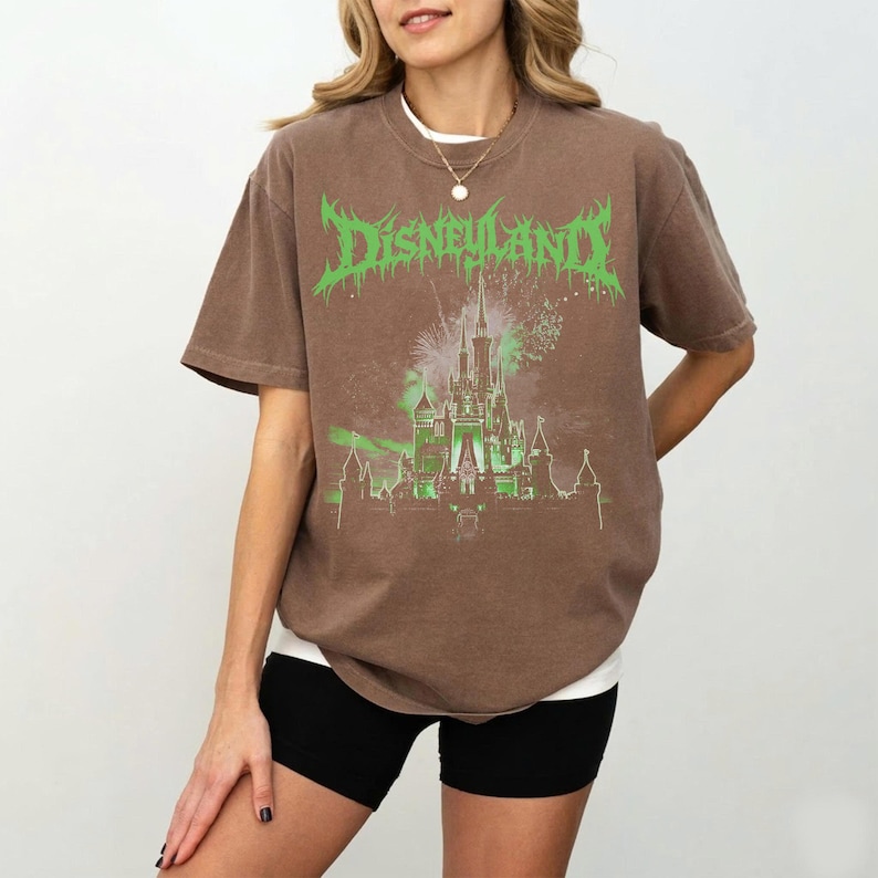 Puede incluir: Camiseta marr&oacute;n con la palabra "Disneyland" en texto verde estilizado. La camiseta presenta un gr&aacute;fico verde de un castillo con fuegos artificiales. La camiseta se usa con pantalones cortos negros y una camiseta interior blanca.