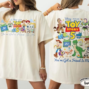 Może przedstawiać: Kremowa koszulka z kolorową grafiką Toy Story, przedstawiającą postacie takie jak Woody i Buzz Lightyear. Prz&oacute;d i tył koszulki przedstawiają logo Toy Story 5 i frazę "You've Got a Friend In Me."