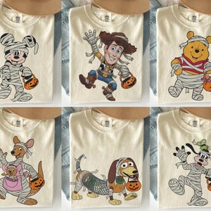 May include: A collection of six cream-colored t-shirts, each featuring a different cartoon character wrapped in mummy bandages and holding a jack-o'-lantern candy bucket. Characters include Mickey Mouse, Woody, Winnie the Pooh, Kanga with Roo, Slinky Dog, and Goofy.