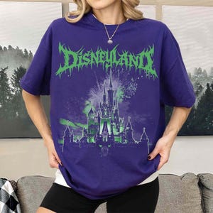 Puede incluir: Una camiseta morada con la palabra "Disneyland" en verde, con una fuente g&oacute;tica. Debajo, una ilustraci&oacute;n verde y blanca de un castillo con fuegos artificiales. La camiseta es de manga corta y de estilo casual.