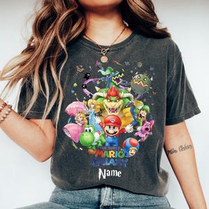 Puede incluir: Camiseta gris oscuro con un gr&aacute;fico colorido de personajes del juego Super Mario Galaxy. El dise&ntilde;o incluye a Mario, Bowser, la Princesa Peach y otros personajes. El texto "THE SUPER MARIO GALAXY" y "Name" tambi&eacute;n son visibles.