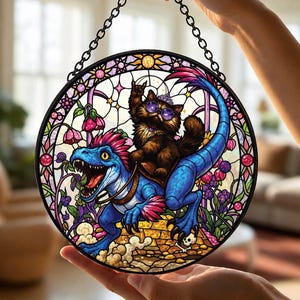 May include: A circular stained glass suncatcher featuring a brown cat wearing purple sunglasses and a crown, riding a blue dinosaur. The design includes pink flowers, a yellow brick path, and a black metal chain for hanging, all framed in black.