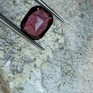 K&ouml;nnte beinhalten: Ein facettierter, rechteckiger Edelstein, der von einer Pinzette gehalten wird. Der Edelstein hat eine tiefrot-violette Farbe mit einer reflektierenden Oberfl&auml;che. Die Pinzette ist silberfarben und der Hintergrund ist eine strukturierte, hellgraue Oberfl&auml;che.
