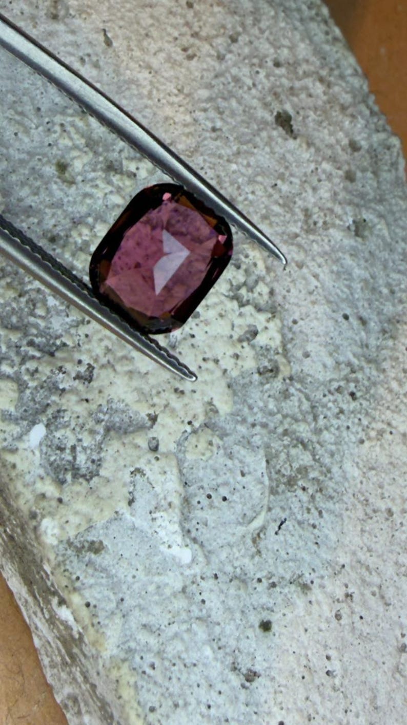 K&ouml;nnte beinhalten: Ein facettierter, rechteckiger Edelstein, der von einer Pinzette gehalten wird. Der Edelstein hat eine tiefrot-violette Farbe mit einer reflektierenden Oberfl&auml;che. Die Pinzette ist silberfarben und der Hintergrund ist eine strukturierte, hellgraue Oberfl&auml;che.