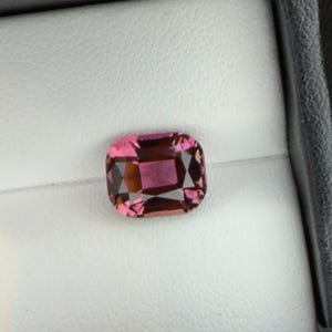 K&ouml;nnte beinhalten: Ein kissenf&ouml;rmiger Edelstein mit einem leuchtenden rosa Farbton und inneren Facetten. Der Edelstein ist vor einem wei&szlig;en Hintergrund in einer schwarzen Box platziert, was seine Farbe und Klarheit hervorhebt. Ideal f&uuml;r Schmuck.