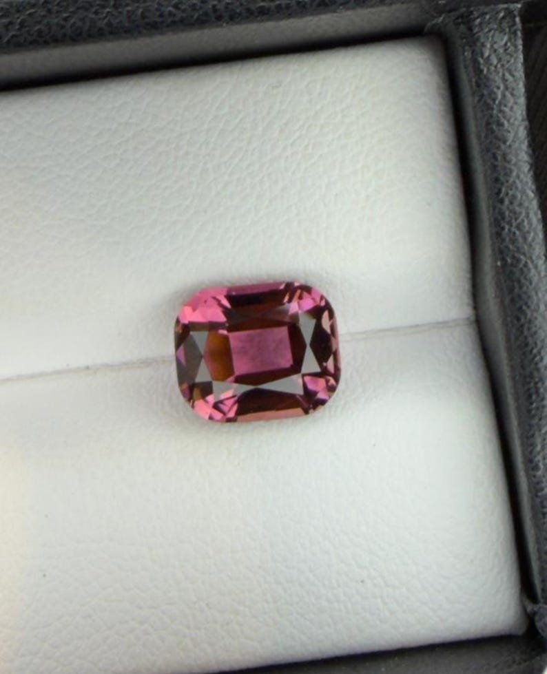 K&ouml;nnte beinhalten: Ein kissenf&ouml;rmiger Edelstein mit einem leuchtenden rosa Farbton und inneren Facetten. Der Edelstein ist vor einem wei&szlig;en Hintergrund in einer schwarzen Box platziert, was seine Farbe und Klarheit hervorhebt. Ideal f&uuml;r Schmuck.