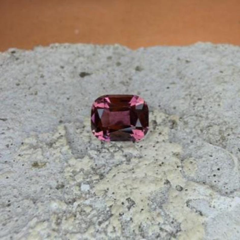 K&ouml;nnte beinhalten: Ein kissenf&ouml;rmiger Edelstein mit einem tiefen, r&ouml;tlich-rosa Farbton. Der Stein liegt auf einer strukturierten, hellgrauen Oberfl&auml;che mit einem kontrastierenden orange-braunen Hintergrund. Die Facetten des Edelsteins reflektieren das Licht.
