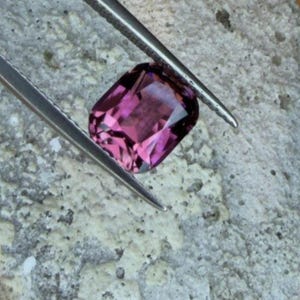 K&ouml;nnte beinhalten: Ein facettierter, rechteckig geschliffener, purpurrosa Edelstein wird von einer silbernen Pinzette gehalten. Der Edelstein liegt auf einer strukturierten, hellgrauen Oberfl&auml;che, die seine Klarheit und Farbe zeigt. Die Pinzette ist angewinkelt, um die Form des Edelsteins hervorzuheben.