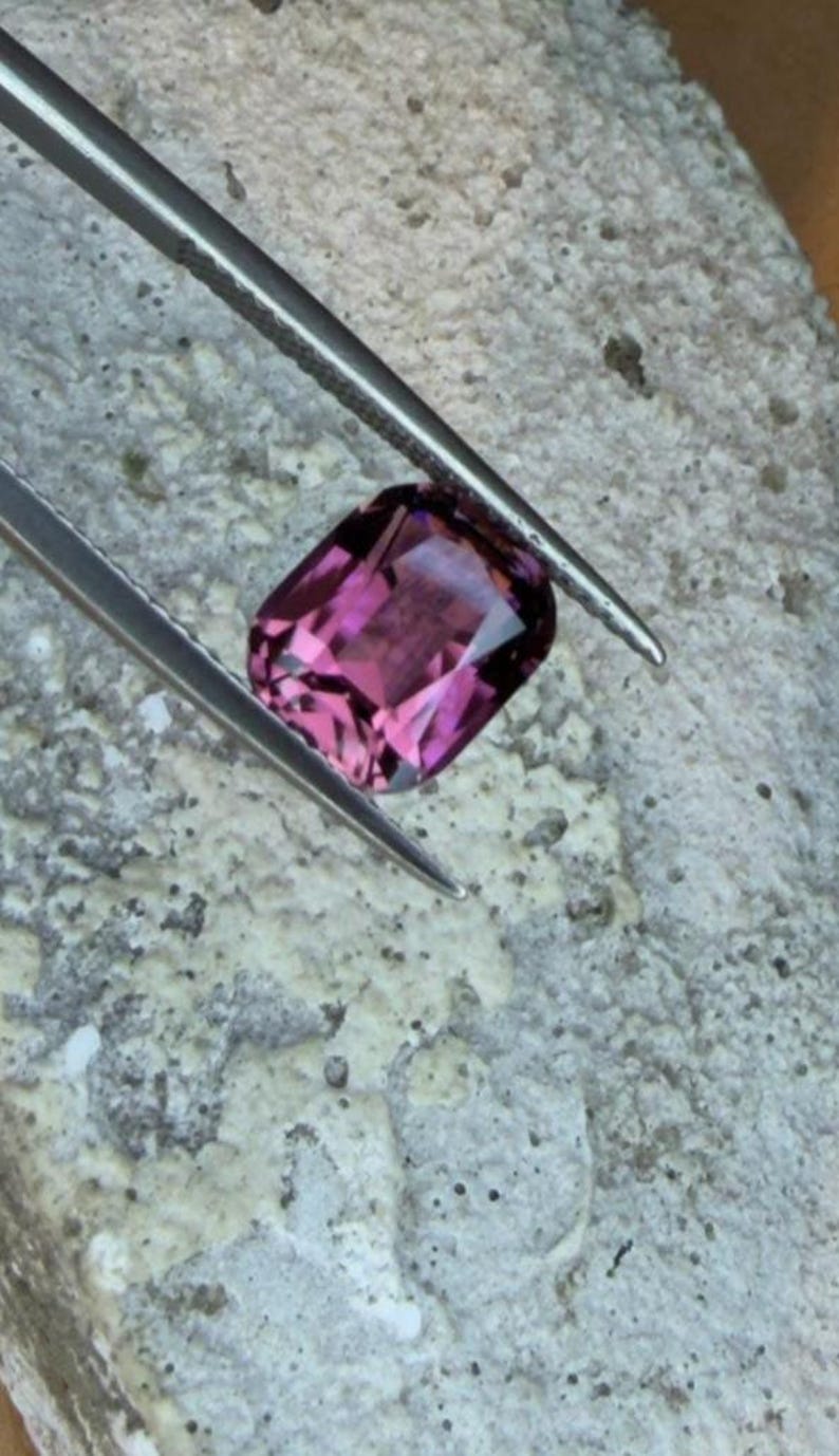 K&ouml;nnte beinhalten: Ein facettierter, rechteckig geschliffener, purpurrosa Edelstein wird von einer silbernen Pinzette gehalten. Der Edelstein liegt auf einer strukturierten, hellgrauen Oberfl&auml;che, die seine Klarheit und Farbe zeigt. Die Pinzette ist angewinkelt, um die Form des Edelsteins hervorzuheben.