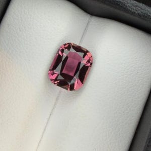 K&ouml;nnte beinhalten: Ein facettierter, rechteckiger Edelstein in tiefem Rosa, eingebettet in einen wei&szlig;en Hintergrund in einer schwarzen Box. Der Schliff des Steins zeigt innere Facetten, die seine Brillanz und Farbe verst&auml;rken.
