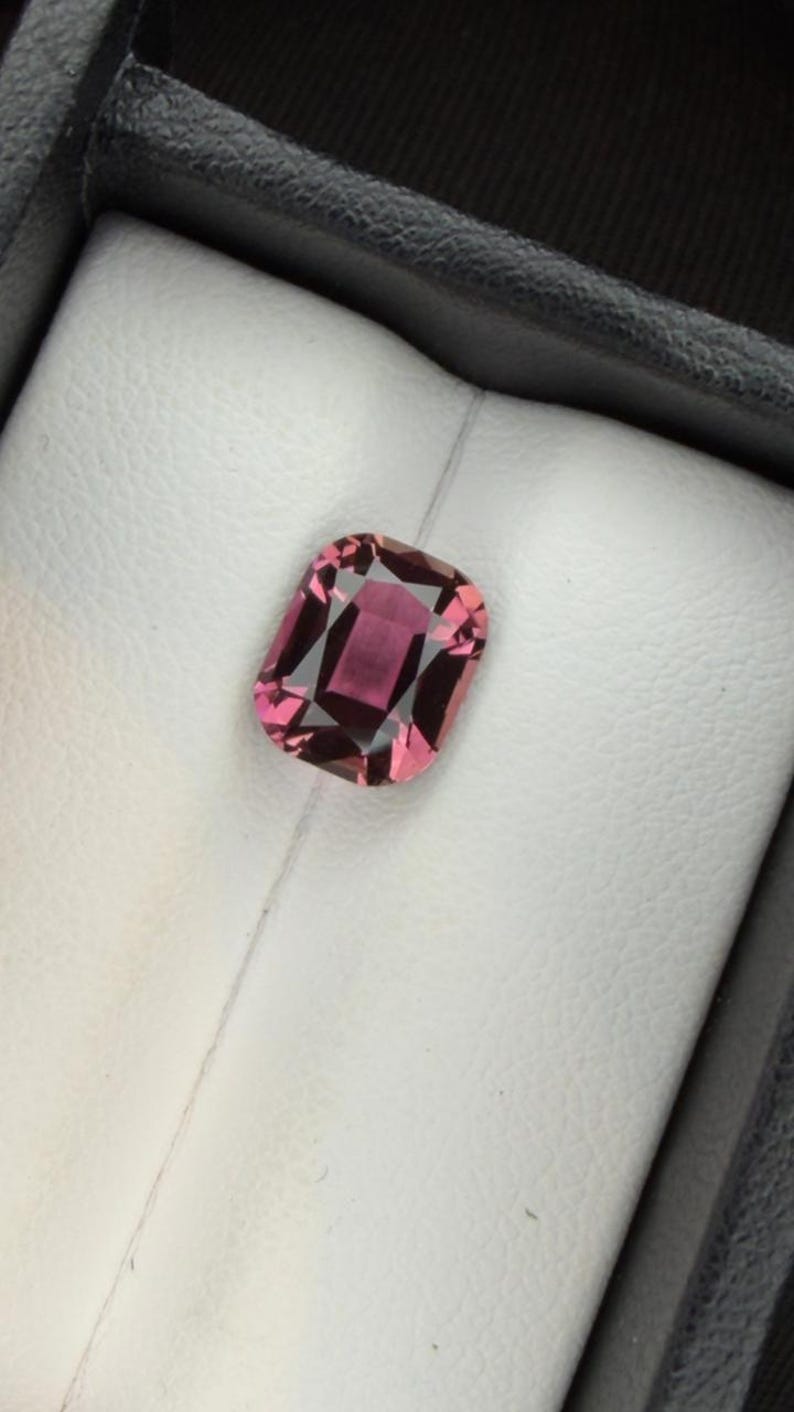 K&ouml;nnte beinhalten: Ein facettierter, rechteckiger Edelstein in tiefem Rosa, eingebettet in einen wei&szlig;en Hintergrund in einer schwarzen Box. Der Schliff des Steins zeigt innere Facetten, die seine Brillanz und Farbe verst&auml;rken.
