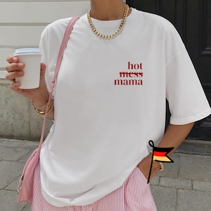 Könnte beinhalten: Weißes T-Shirt mit dem roten Text "hot mess mama". Das Outfit wird mit einer rosa-weiß gestreiften Hose und einer rosa Umhängetasche kombiniert. Eine goldene Halskette und Armbänder sind ebenfalls zu sehen.
