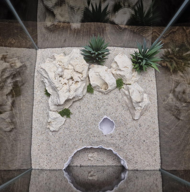 May include: A terrarium setup with a light-colored sand substrate, white rocks, and green artificial plants. The terrarium has a cave-like opening and a small hole. The background features a textured, brown surface.