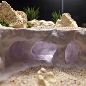 May include: A miniature cave diorama with a sandy foreground and textured, off-white rock formations. The cave interior glows with a soft purple light, and green plants are visible in the background. The scene evokes a natural, desert-like environment.