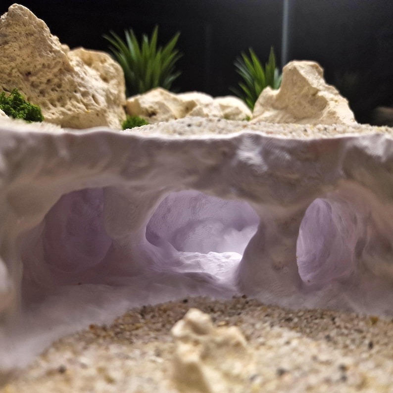 May include: A miniature cave diorama with a sandy foreground and textured, off-white rock formations. The cave interior glows with a soft purple light, and green plants are visible in the background. The scene evokes a natural, desert-like environment.