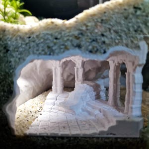 May include: A white, miniature, ancient-style ruin with columns and a pathway, designed for an aquarium. The structure is nestled within a sandy, textured environment, creating a detailed underwater scene. The design includes steps and archways.