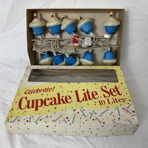 Pode incluir: Uma sequência de 10 luzes em forma de cupcake numa caixa. Os cupcakes têm cobertura branca, invólucros azuis e velas amarelas. A caixa diz "Celebrate! Cupcake Lite Set".