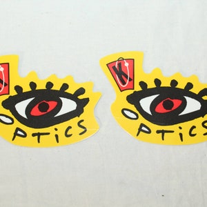 May include: Two yellow die-cut stickers with a stylized eye design. The eye is black and white with a red iris. Above the eye is a red and white graphic. The word "OPTICS" is printed in black below the eye.
